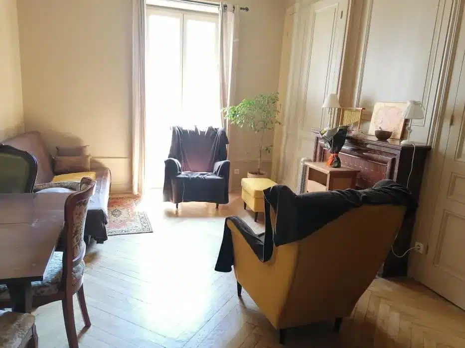 Salon chaleureux avec fauteuil jaune, parquet en chevrons, cheminée ancienne et lumière naturelle par la fenêtre.