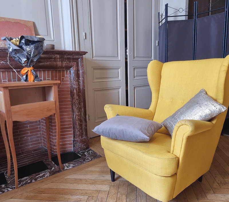 Fauteuil jaune avec coussins gris et argenté près d'une cheminée ancienne et d'un guéridon en bois dans un salon.